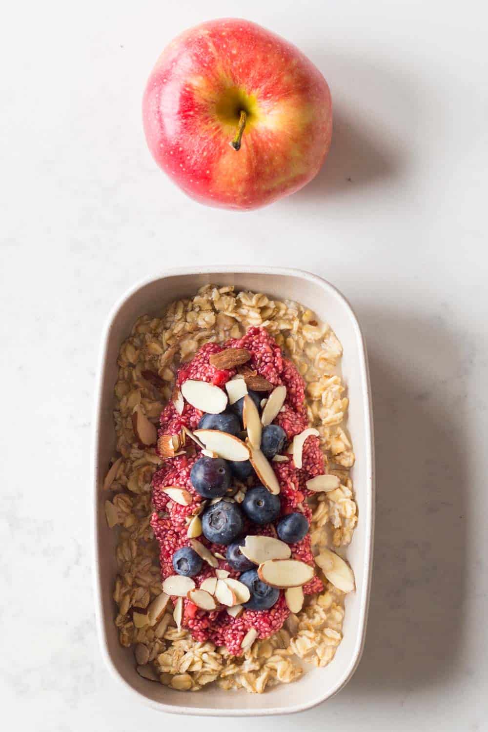 Meal prep container filled with overnight, chia pudding jam, sliced almonds, blueberries and, an apple on the side.